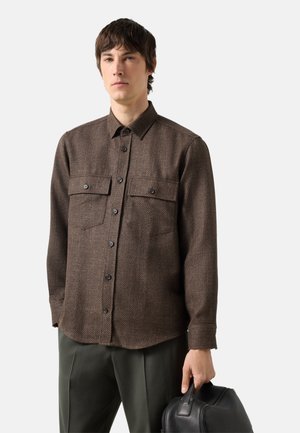 Mann trägt ein braunes Hemd mit Knöpfen und zwei Brusttaschen, dunkle Hose, hält eine schwarze Lederreisetasche vor schlichten Hintergrund.