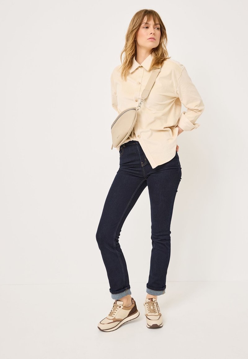 Camicia button-up beige chiaro, jeans skinny blu scuro e sneakers beige con accenti marroni. Design semplice e forma aderente in tutto.