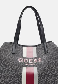 Czarna torba tote o fakturze, z dwoma czarnymi uchwytami, z centralnym paskowanym wzorem w kolorze czerwonym i białym oraz wyraźnie wyeksponowanym napisem "GUESS".