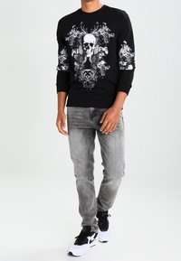 Felpa nera con stampa di teschio e fiori bianchi, abbinata a jeans grigi e sneakers in bianco e nero. Texture liscia, collo a giro.
