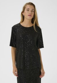 Vrouw met lang blond haar, gekleed in een zwart top met korte mouwen en een rok bedekt met kleine glanzende zwarte pailletten, staand tegen een effen achtergrond.