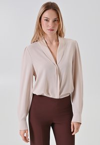 Hellbeige Bluse mit langen Ärmeln und tiefem V-Ausschnitt aus weichem Stoff, kombiniert mit dunkelbraunen, hoch taillierten Hosen. Sanfte Textur, lässige Passform.