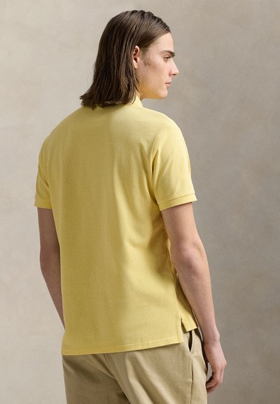 Muškarac s smeđom kosom do ramena, odjeven u žuti polo s kratkim rukavima i bež hlače, okrenut leđima prema jednobojnoj pozadini.