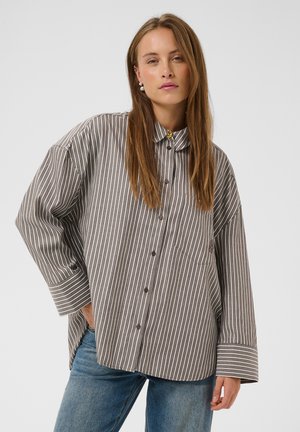 Femme portant une chemise ample à rayures verticales grises et blanches boutonnée et un jean bleu, debout devant un fond uni.