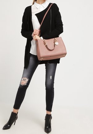 Kvinde iført sort cardigan, hvid skjorte, revnede mørke skinny jeans, sorte ankelstøvler med hæl, holder en lyserød håndtaske med gyldne kædedetaljer.