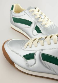 Baskets en cuir argenté avec des accents verts, des lacets crème et une semelle en caoutchouc gomme. Présente un design élégant et des détails de texture.