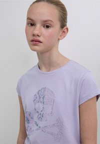 T-shirt in cotone lilla con maniche a cappuccio, caratterizzata da un design grafico di teschio e tibie incrociate in colori pastello e dettagli testuali.