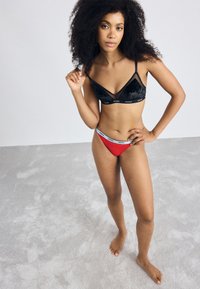 Black velvet bralette with adjustable straps, featuring a geometric design. Red high-cut thong with white branded waistband, displayed on a light background.