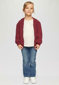 Maronowy hoodie na zamek, biały t-shirt z wzorem serc, niebieskie dżinsy w pionowe paski oraz białe trampki. Prosta, codzienna stylizacja.