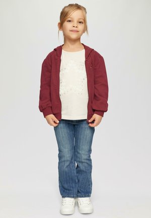 Maronowy hoodie na zamek, biały t-shirt z wzorem serc, niebieskie dżinsy w pionowe paski oraz białe trampki. Prosta, codzienna stylizacja.