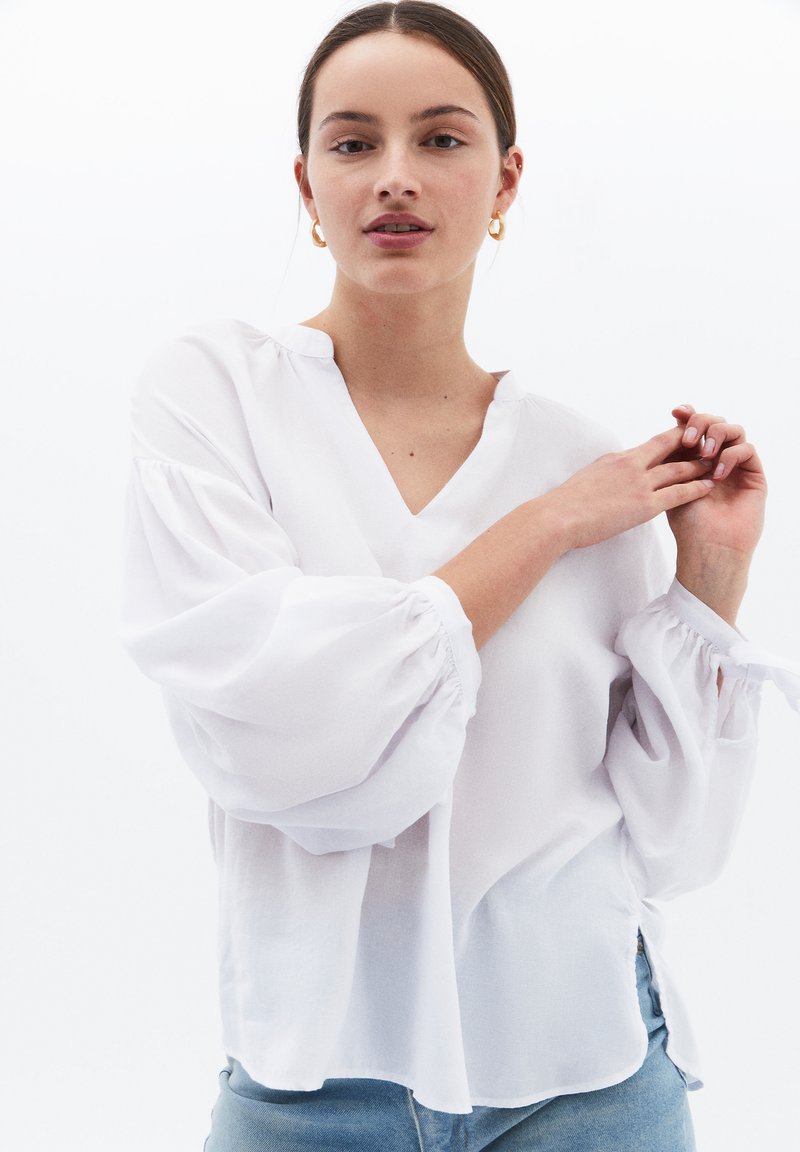 Blusa bianca a maniche lunghe con scollatura a V, maniche a palloncino arricciate e spacchi laterali; realizzata in tessuto leggero con una consistenza morbida.