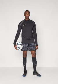 Svart langermet topp med grå detaljer, glidelås krage; mørke mønstrede shorts; svarte sokker; svarte fotballsko; holder en mønstret hvit fotball.