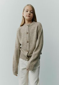 Beige gestrickte Strickjacke mit rundem Ausschnitt, durchgehender Knopfleiste und gerippten Bündchen, kombiniert mit weißen Hosen mit schwarzen Punkten.