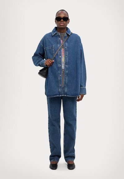 Jeans skjorte og bukser i middelblå, med en avslappet passform og knapp-lukking foran. Tilbehør inkluderer svarte solbriller og en liten veske.