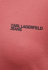 Rød bomullsstoff med ribbet tekstur og et svart trykt logo som sier "KARL LAGERFELD JEANS" sentrert på den nederste delen.