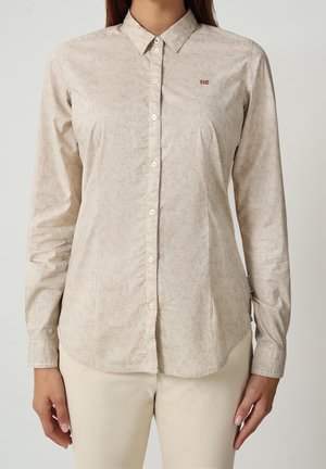 Frau trägt beige Langarm-Bluse mit feinem Kreis-Muster und kleinem rot-weißem Logo auf der Brust, kombiniert mit hellen Hosen.