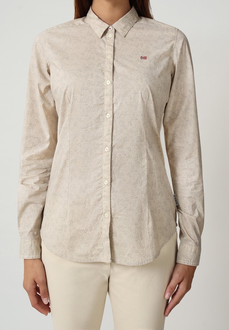 Femme portant une chemise beige à manches longues avec un motif circulaire subtil et un petit logo rouge et blanc sur la poitrine, associée à un pantalon clair.
