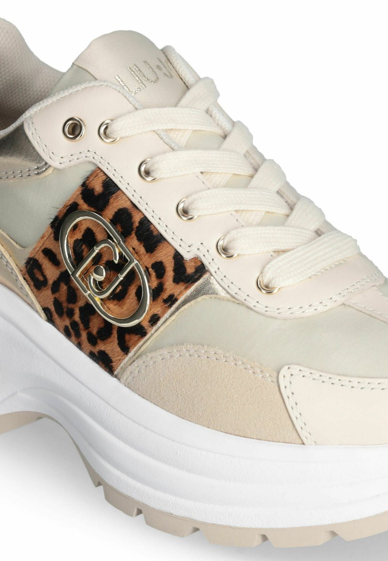 Zapatilla con una parte superior de cuero blanco y beige, que presenta un panel de estampado de leopardo, acentos metálicos y un detalle de logo en el lateral.