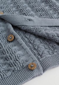 Világoskék kötött kardigán texturált gyémántmintákkal, fa gombokkal és bordázott szegéllyel, részben kigombolva fehér háttéren.