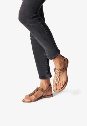 Brune flettede sandaler med vævet remdesign, flad sål og justerbar ankelrem, båret sammen med mørke jeans.