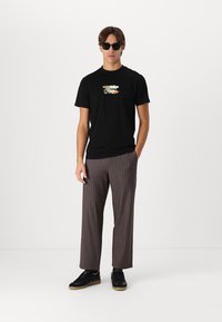 Čierne bavlnené tričko s farebným grafickým dizajnom, skombinované s hnedými pruhovanými širokými nohavicami. Čierne tenisky a slnečné okuliare dotvárajú celkový vzhľad.