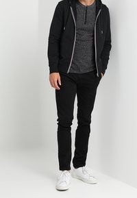 Haut à capuche zippé noir avec cordons de serrage blancs, chemise henley grise, pantalon slim noir et baskets blanches. Tenue décontractée simple.