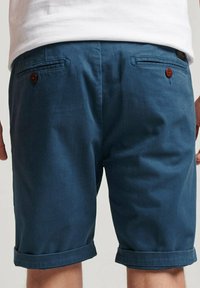 Pantalones cortos azules hechos de una tela ligera, con dobladillos enrollados, dos bolsillos traseros con botones marrones y una textura sutil.