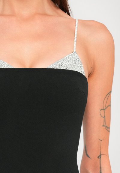 Žena v čiernych bezramienkových šatách vrstvených cez strieborný top s rhinestone špagetovými ramienkami, s minimalistickým lineárnym tetovaním na hornej časti ramena.