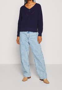 Person klädd i en marinblå V-ringad virkig tröja, ljusblå vida jeans och beiga strukturerade ankelskor som står mot en enfärgad bakgrund.