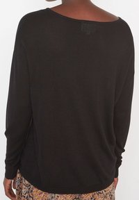 Haut noir à manches longues avec une coupe ample, une texture côtelée, un large décolleté et des fentes sur les côtés à l'ourlet, porté avec un tissu à motifs en dessous.