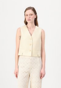 Cropped beige vest, featuring a V-neck, three buttons, and side pockets, paired with light lace-patterned trousers.