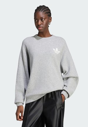 Ung kvinde med flettet hår iført en lysegrå Adidas-trøje og sorte bukser, stående med den ene hånd i lommen.