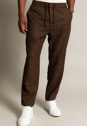 Man draagt bruine geruite broek met trekkoord en zijzakken, en witte sneakers, staande tegen een effen lichte achtergrond.