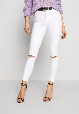 Vrouw staand, jurk met gescheurde witte skinny jeans, zwarte gespikkelde riem, paarse gebreide trui en zwarte sandalen met bandjes en hoge hakken.