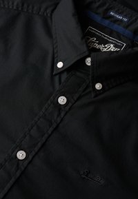 Sort button-up skjorte i regular fit. Har en glat tekstur, hvide knapper og et diskret logo på kanten. Klassisk kravedesign.