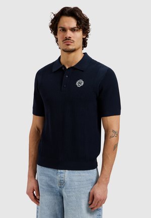 Man draagt een marineblauw gebreid poloshirt met korte mouwen en een wit geborduurd logo, gecombineerd met lichtblauwe jeans, staand tegen een effen achtergrond.