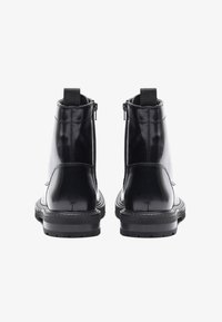 Par de botines de cuero negro brillante con tiradores y cremalleras laterales, vistos desde atrás sobre un fondo blanco.