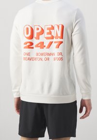 Sudadera blanca con texto naranja en negrita: "ABIERTO 24/7" con detalles de dirección. Puños y dobladillo acanalados, textura suave, corte relajado. Pantalones cortos negros visibles.