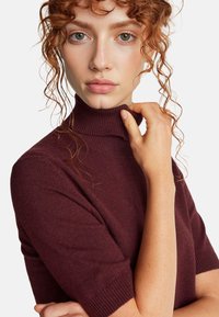 Frau mit lockigem rotem Haar, die einen burgunderfarbenen Rollkragenpullover trägt und mit einer Hand den Kragen hält, neutrale Miene, weißer Hintergrund.