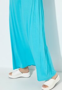 Ein langes, fließendes türkisfarbenes Kleid mit glatter Textur, kombiniert mit weißen Plattform-Slidesandalen mit offenem Zeh und abgerundeten Sohlen.