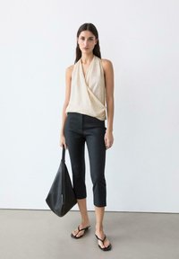 Frau in beigeärmligem, drapiertem Oberteil, schwarzen, knöchellangen Hosen, schwarzen Flip-Flops, hält eine große schwarze Tragetasche, steht vor einer weißen Wand.