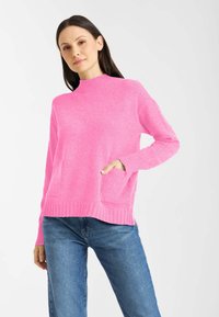 Różowy sweter z miękkiego materiału, z wysokim kołnierzem, prążkowanym wykończeniem i dwoma przednimi kieszeniami. Zestawiony z niebieskimi dżinsami.