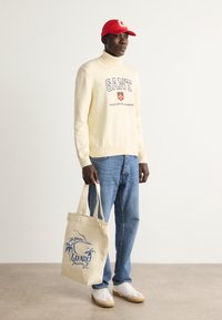Cremefarbener Pullover mit "GANT" Logo, blaue Jeans, weiße Sneakers mit beige Sohle und eine beigefarbene Tragetasche mit blauen Palmenmotiven. Rote Kappe.