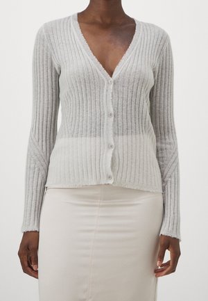 Osoba o ciemnej karnacji ubrana w jasnoszary prążkowany kardigan na guziki z dekoltem w serek oraz kremową spódnicę na jednolitym tle.