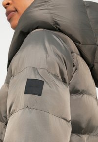 Doudoune grise avec une texture lisse, dotée d'un col montant et d'un patch logo noir sur la manche gauche.