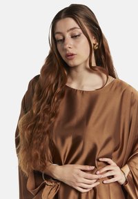 Top in satin marrone con un design fluido e ampio scollo. Capelli lunghi e mossi, bracciale in argento minimale al polso.