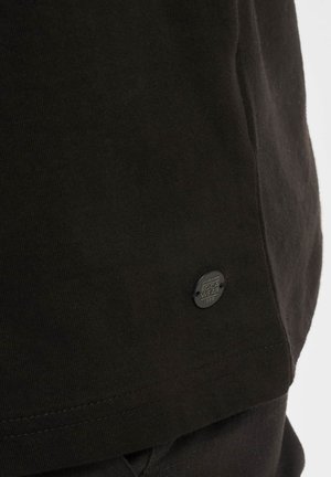 Mustmustärgne puuvillane T-särk, millel on ribadega kaelus ja külgõmblus. Särgil on hemme lähedal väike must Roca Wear logo silt.