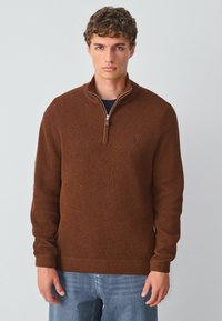 Brązowy sweter dzianinowy z kołnierzem na zamek błyskawiczny do połowy. Ma fakturowany materiał oraz ściągacze przy mankietach i dolnym hemie. Noszony na granatowej koszuli i niebieskich dżinsach.