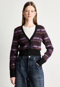 Crni kardigan s višebojnim uzorcima, s natpisom "JEANS TOMMY". Dizajn s V-izrezom, kopčanje na gumbi i rebrasti manžeti. Nosiv preko bijele košulje.