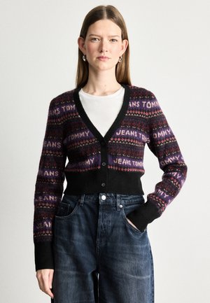 Juodas kardiganas su daugiaspalviais raštais, su užrašu "JEANS TOMMY." V-poškas dizainas, mygtukų uždarymas ir raibos rankogaliai. Dėvimas ant baltos marškinėlių.